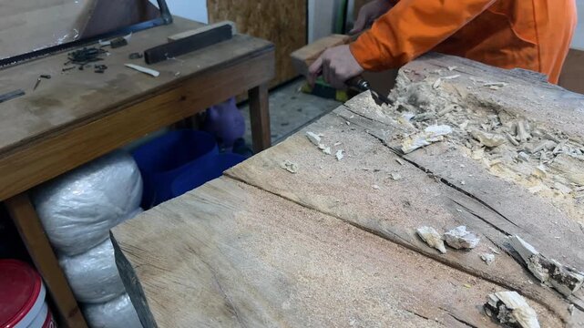 A carpenter in overalls chiselling a wooden slab in a woodworking shop. A person working. Tools, chisels, and gouges. Woodworking. Sawdust, wood chips. Real people. Lifestyle, Authentic, Candid