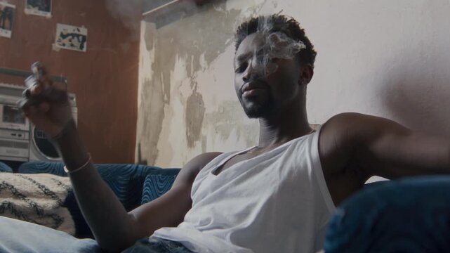Portrait of young Black man smoking cigarette and exhaling smoke, looking at camera confidently as sitting on couch in modest worn apartment with peeling wall and vintage stereo in background
