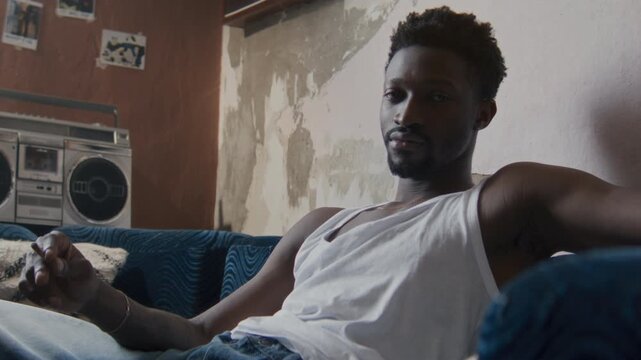 Portrait of young African American man sitting on couch in modest room with vintage boombox and peeling wall in background, holding cigarette and looking seriously at camera