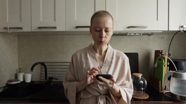 chemo patient with shaved head scrolls and types on smartphone in the kitchen, quiet self care moment at home, managing treatment side effects and chatting with support group