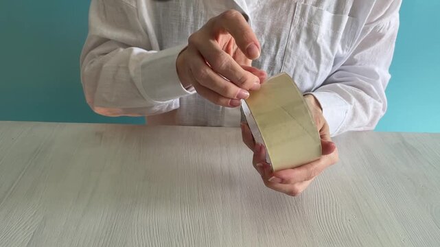 close-up of hands peeling and tearing transparent packing tape from a roll on a wooden tabletop with sharp ripping sounds, concept of packaging, shipping, diy repair, office work