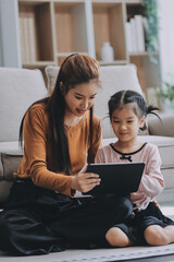 Fototapeta premium A cheerful Asian mother helps her young daughter do homework and color a book at home, while the father works on a laptop in the background. A cozy, bright, and modern family lifestyle.