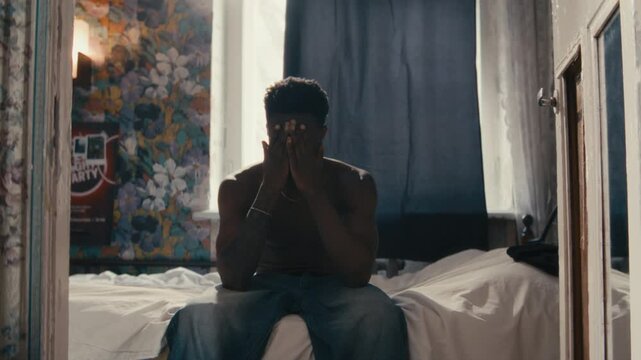 Young shirtless Black man sitting on bed in softly lit bedroom, rubbing his face and staring pensively ahead, then getting up and leaving. Zoom in shot