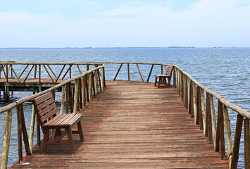 Obraz premium IGUABA GRANDE, RIO DE JANEIRO, BRAZIL – January 18, 2025: A rustic wooden deck extends over the tranquil waters of the Araruama Lagoon in Iguaba Grande, Rio de Janeiro, where two benchs face the calm 