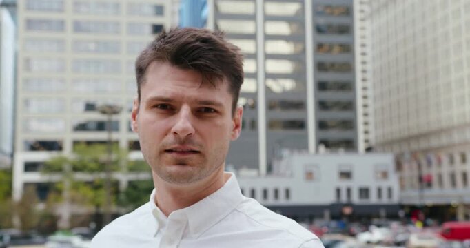 A close-up shot of a middle-aged man in a white shirt standing focused and looking into the camera among the tall buildings of a business center in a big city.