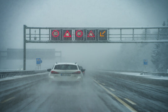 A car drives on the snow-covered A8 motorway under variable traffic signs indicating speed limits and warnings of slippery roads while it is snowing heavily