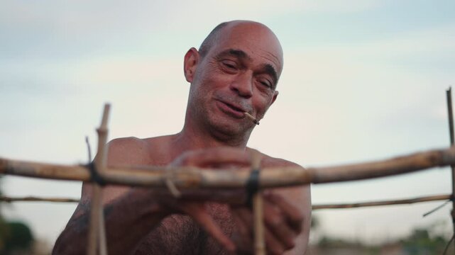 Happy shirtless gardener with a cigarette smiling while tying some reeds for his urban garden
