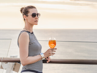 Fashionable woman with a glass of drink standing on the empty deck of a cruise ship against the backdrop of sea waves. Sunny day. Chill lifestyle, luxury travel. Perfect for holiday and travel themes © Svetlana