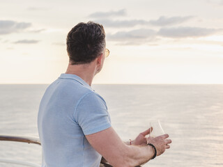 Stylish man with a glass of drink standing on the empty deck of a cruise ship against the backdrop of sea waves. Sunny day. Chill lifestyle, luxury travel. Perfect for holiday and travel themes © Svetlana