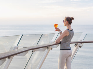 Fashionable woman with a glass of drink standing on the empty deck of a cruise ship against the backdrop of sea waves. Sunny day. Chill lifestyle, luxury travel. Perfect for holiday and travel themes © Svetlana