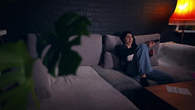 Young Caucasian woman relaxes at home on evening holding remote control and watching television. Slowmo of comfortable mood as she flips through channels. Theme of entertainment and the everyday life.