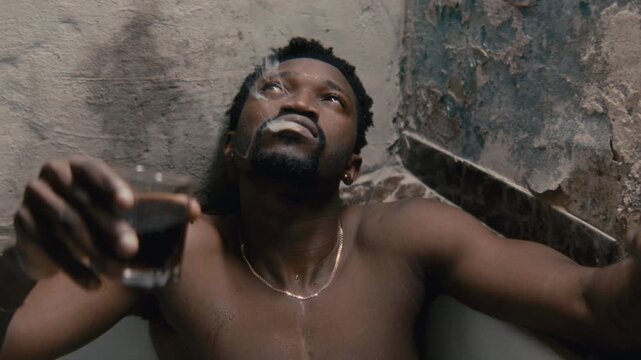 Young African American man relaxing in bathtub against worn textured wall, drinking alcoholic beverage from glass and smoking joint, looking upward deep in thought