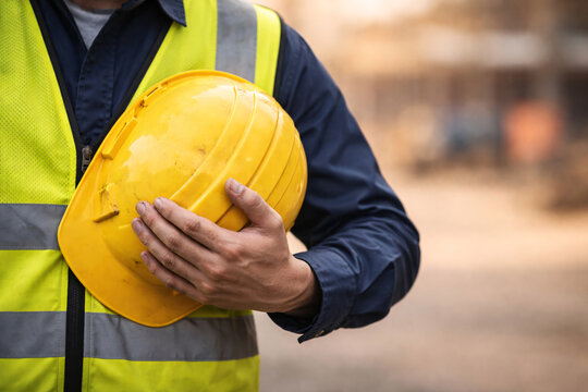 Construction worker in yellow reflective vest holding a yellow hard hat with a blurred background