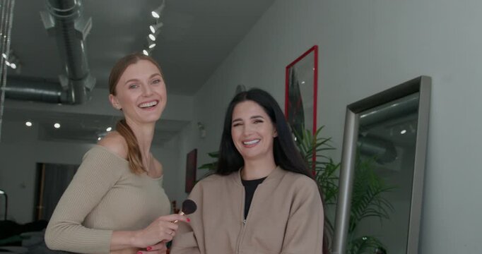 A makeup artist and her client are happily looking at the camera, posing and smiling in a classy salon.