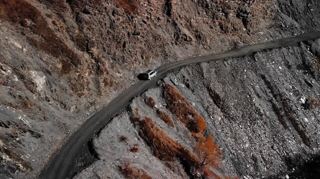 Drone view SUV climbing rugged cliffs on narrow mountain road in Georgia, white SUV navigating steep switchback above deep ravine, autumn shrubs and rocky slopes, remote geological survey training,