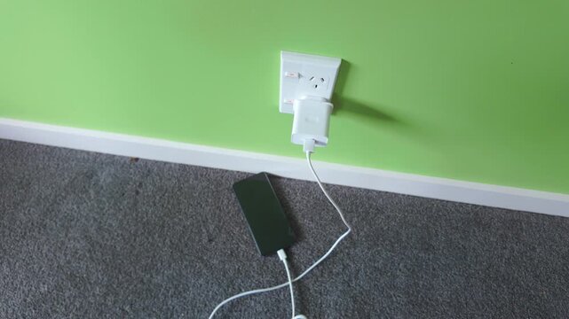 Close-up of a person's hand unplugging several chargers from a power adapter connected to a wall socket. A smartphone remains charging on the floor