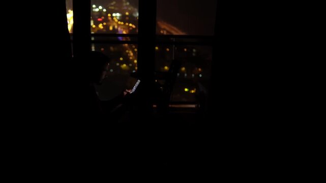 nighttime window silhouette using phone, latenight freelancer checking messages, city lights shimmer outside, dim screen glow illuminates hands and partial face, mood quiet and introspective, compact