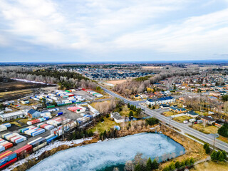 Fototapeta premium Industrial storage yard with rows of shipping containers by a frozen pond and road, bordering houses and new apartment complexes in barrie, ontario, showing urban growth and infrastructure
