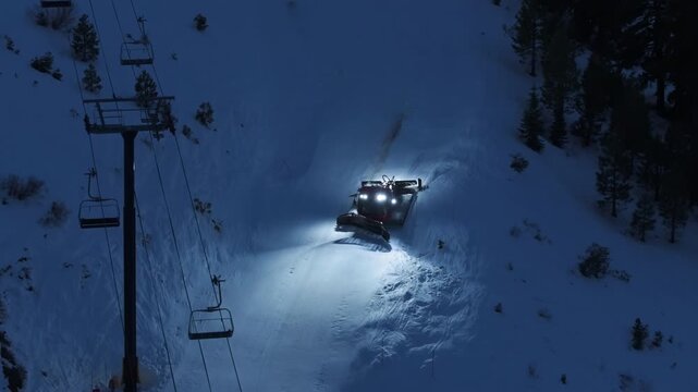 Aerial close up of a snowcat grooming a ski run at night. Drone footage shows tracked vehicle smoothing fresh snow for winter sports at mountain resort.