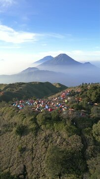 Vertical drone footage over Mount Prau savanna in Wonosobo. Camp tents scattered across hills with Mount Sindoro and Mount Sumbing dominating horizon.