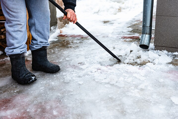 Worker on roof with crowbar breaking and removing ice covered sidewalk formed by melting water from drain pipe during winter thaw.
