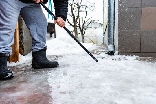 Worker on roof with crowbar breaking and removing ice covered sidewalk formed by melting water from drain pipe during winter thaw.