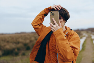 Fototapeta premium Young woman lifestyle blogger in orange jacket takes selfie with smartphone outdoors on rural path. Casual autumn outfit and natural background create relaxed, modern vibe.