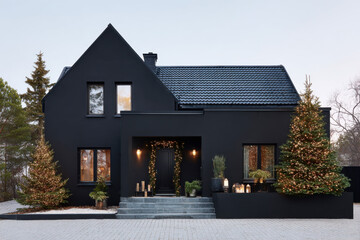A matte black modern house sits in a snowy landscape decorated with several small Christmas trees