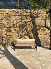 Empty wooden park bench sits against a textured stone wall with dappled sunlight and tree shadows
