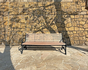 Empty wooden park bench sits against a textured stone wall with dappled sunlight and tree shadows