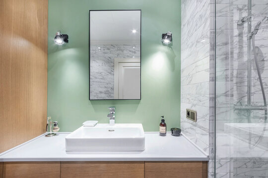 A bathroom is set up with a square sink on a light counter. Above the sink is a rectangular mirror. The walls are painted green, and a shower area is visible nearby