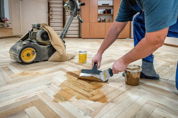 Worker in blue fills the gaps cracks of the old wooden parquet with a spatula after sanding. Floor renovation