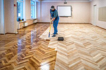 Worker in blue overalls uses a roller to coating floors. Lacquering wooden parquet floors.