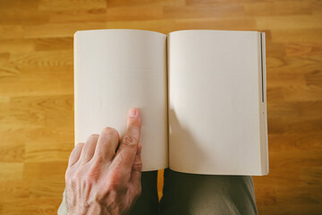POV of person holding open blank book mockup with finger pointing at empty page over wooden floor background.