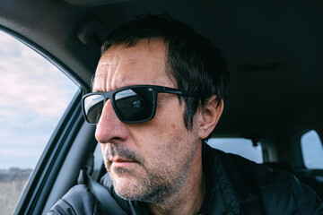 Middle aged man wearing sunglasses while driving in car, seat belt fastened, serious focused expression, realistic lifestyle portrait inside vehicle.