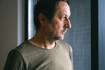 Mature adult man with unkempt hair and facial stubble looking out the window of his apartment, sad and disappointed expression, quiet reflective indoor moment.