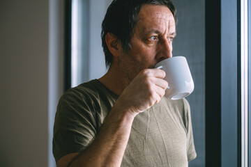 Middle aged man drinking coffee from a cup while standing by a window, thoughtful mood, natural daylight, quiet indoor lifestyle moment.