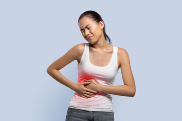 A woman stands in a simple indoor space and holds her stomach with a pained expression. She seems to be experiencing discomfort. The background is light blue.