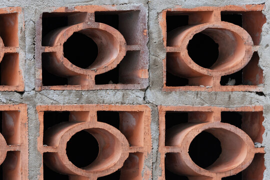 A close-up of a terracotta cobogo wall, showing a geometric pattern of circular and rectangular openings. Rough reddish-brown clay blocks and grey mortar. 