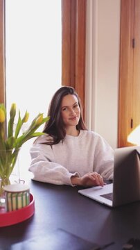 Happy young woman working on laptop at home office, smiling female freelancer typing on keyboard and looking at screen with satisfaction, positive brunette girl enjoying remote work in cozy room