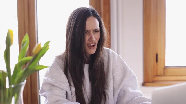 Horizontal video of a young brunette woman in a white sweatshirt working on her laptop at home, angrily waving her hands and reacting to bad news, with yellow tulips in a vase visible on the table.