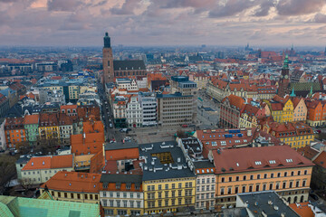 Naklejka premium Aerial dusk view of Wroclaw, Poland, showing Market Square, St. Elizabeth's brick tower, Old Town Hall clock tower, pastel facades, red roofs, and a cloud streaked sky.