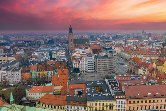 A sweeping aerial view shows Wroclaw, Poland, Old Town at sunset, with St. Elizabeth's Church, the Rynek, red tiled rooftops, modern blocks, and pedestrians moving.