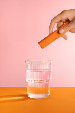 A hand dissolving an orange stick powder in water on pink background.