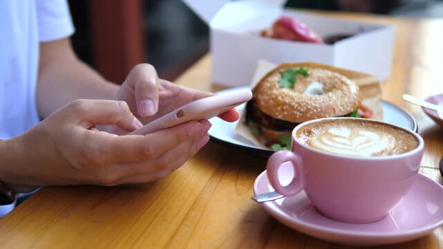 Closeup of female hands scrolling and typing on a modern smartphone while sitting at a wooden table in a cafe with a pink cup of cappuccino, latte art, and a fresh bagel for breakfast