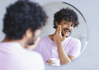 A man stands in front of a mirror and applies skin cream to his face. He has curly hair and wears a light shirt. © Prostock-studio