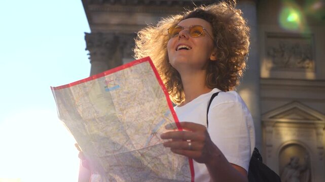 Beautiful happy woman traveler with a backpack holding a paper map while enjoying the sights of a historic european city at sunset with beautiful sun flare and a smile on her face