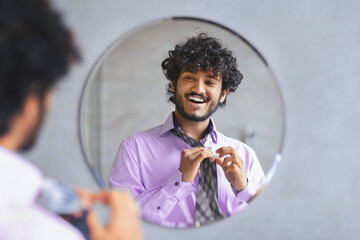 A man looks at his reflection in a round mirror while he adjusts his tie. He wears a light purple shirt and has curly hair. The setting appears tidy and well-lit with a simple background. © Prostock-studio