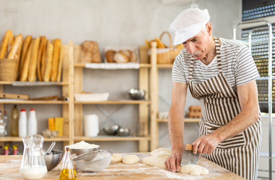 Elderly man baker cutting dough with scraper