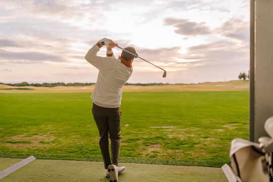 Male golfer in white long-sleeve shirt swings golf club on driving range with green grass and cloudy sky in the background during late afternoon